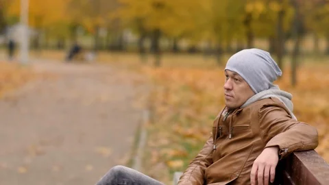 Attractive guy in a gray hat is resting while sitting on a bench in the autumn Stock Footage 120185631