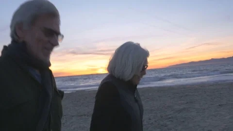 Attractive happy smiling mature 60s couple walking on beach at sunset Stock Footage 74434026