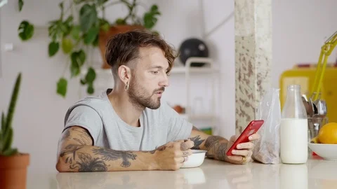 Attractive Hispanic guy eating breakfast flakes while using red mobile phone Stock Footage 108795637