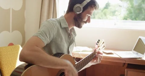 Attractive hispanic man sitting at window learning to play guitar using laptop Stock Footage 73369602
