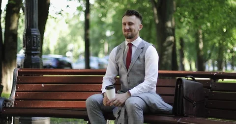 An attractive man with a beard is sitting on a bench in the park and drinking a Stock Footage 116049204