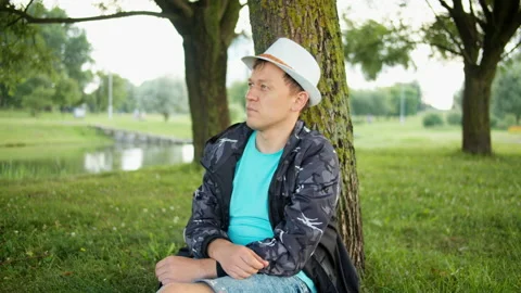 Attractive man in hat resting under a tree in a city park, tracking camera Stock Footage 139141522