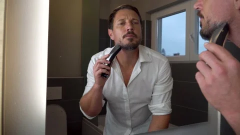 An attractive man looking himself in the mirror while shaving his beard. 4K Stock Footage 139885564