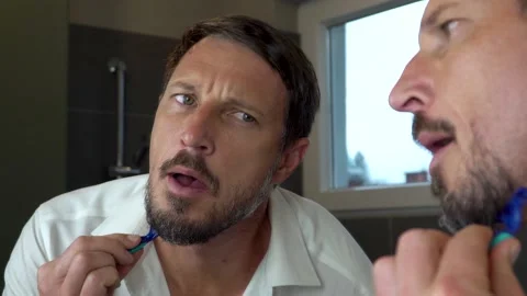 An attractive man looking himself in the mirror while shaving his beard. 4K Stock Footage 139885636