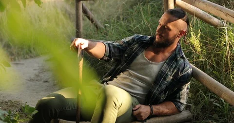 An attractive man in plain clothes sits on a path leaning against a wooden raft Stock Footage 126497565