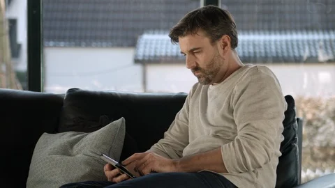 Attractive man sitting on a couch with his ipad  is checking his E-mails Stock Footage 87568411