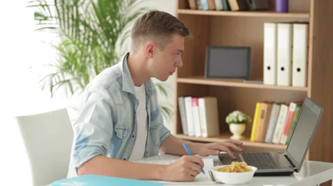 Attractive man student sitting at table using laptop and eating chips Stock Footage 27692044