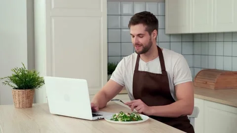 An attractive man uses a laptop while sitting in the kitchen and eats a vegan Stock Footage 151403903