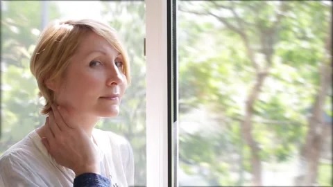 An attractive mature woman stands at the picture window admiring nature Stock Footage
