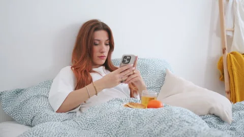 Attractive redhead having breakfast in bed, uses smartphone, front view Stock Footage 306679296