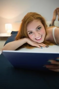 Attractive redhead using digital tablet lying on her bed smiling at camera Stock Photos
