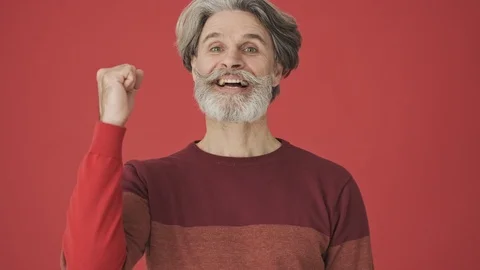 Attractive satisfied gray-haired bearded man doing winner gesture over red wall Stock Footage 120783097