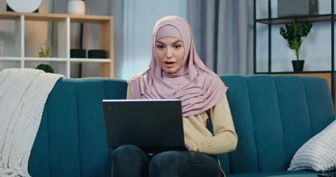 Attractive satisfied smiling young arabic woman in hijab sitting on the couch at Stock Footage 150113778