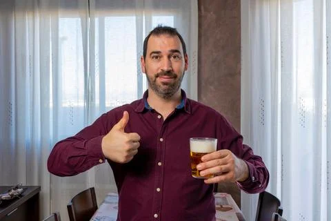 Attractive smiling bearded man in a purple shirt with his thumb up and a beer Stock Photos