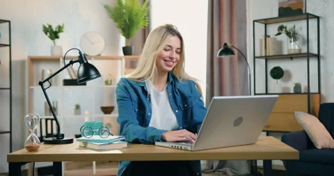 Attractive smiling modern blond young woman sitting at the desk at home and Stock Footage 158449487