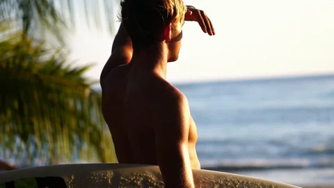 Attractive surfer man looking out towards ocean with surfboard during sunset Stock Footage 108568765