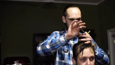 Attractive young barber is cutting human hair with the scissors. He is looking Stock Footage 69964669