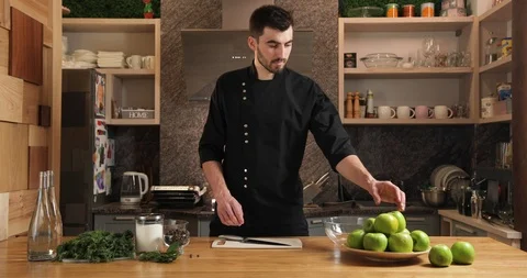 Attractive young chef in a black kettle in the kitchen slices green apples. a Stock Footage 126152901