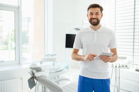 Attractive young dentist in coat using tablet in office and looking at the ca Stock Photos