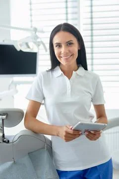 Attractive young dentist in coat using tablet in office and looking at the ca Stock Photos