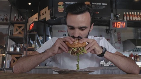 Attractive young guy with a beard bites a burger sitting at a table in a cafe Stock Footage 115634194