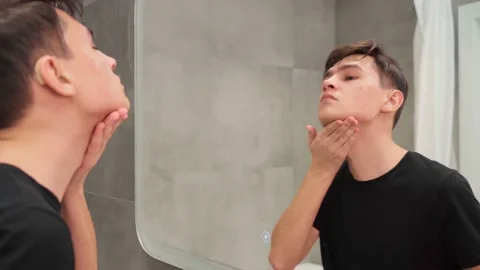Attractive young guy washes his face in the bathroom, looking in the mirror. Guy Video stock 171540334
