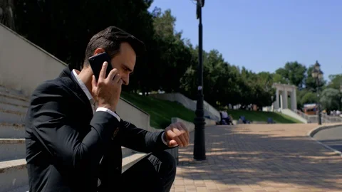 Attractive young man with a beard sitting in a park and talking on the phone. 4k Stock Footage 113923394
