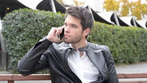 Attractive young man with black jacket and white t-shirt speaks on the phone. Видео 120800260