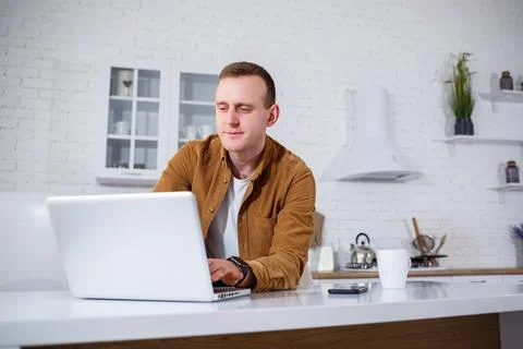 An attractive young man in casual clothes sitting in the kitchen using a lapt 스톡 사진