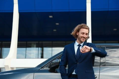 Attractive young man checking time near luxury car outdoors Stock Photos