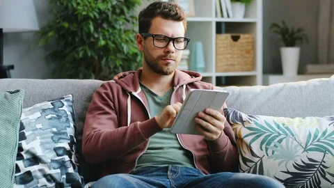 Attractive young man in glasses scrolling and taping on his tablet device while Video stock 83328118