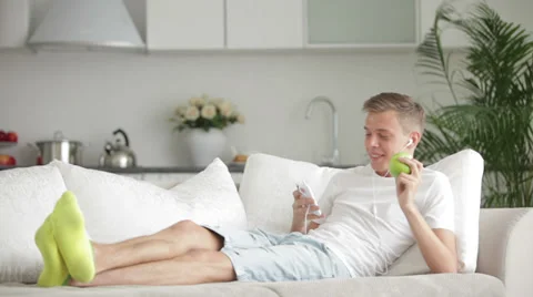 Attractive young man lying on sofa using cell phone and eating apple Stock Footage 27692038