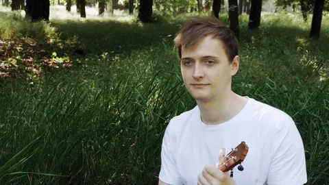 Attractive young man playing a ukulele sitting on grass The man touches the s Stockbeeldmateriaal 108389567