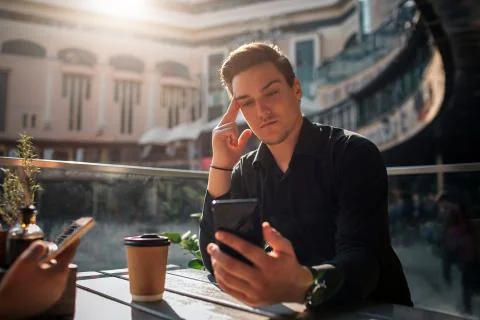 Attractive young man sit at table and look at phoen he hold in hand. Guy is Fotos Stock