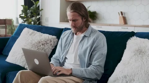 Attractive young man is sitting on a couch at home typing on a laptop looking Stock Footage 195940508