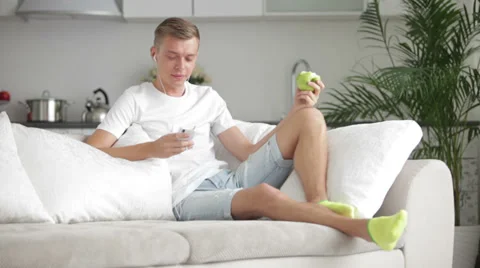 Attractive young man sitting on sofa with cell phone and eating apple Video stock 27692045