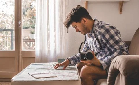 Attractive young man studies a map sitting on the couch. He has the camera an Stock Photos