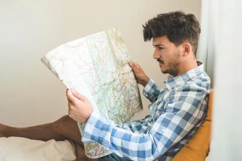 Attractive young man studies a map sitting on his bed. He's thinking about wh Stock Photos