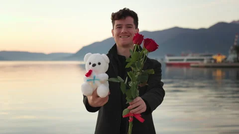 Attractive young man in stylish coat holds out bouquet of red roses and teddy Stock Footage 230970635