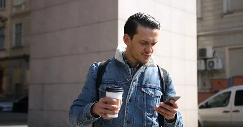 Attractive young man use phone and drinking coffee on the street in sunny spring Stock Footage