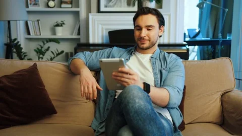 Attractive young man using tablet while relaxing on the sofa at the living room. Stock Footage 77314401