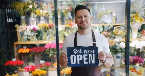Attractive young man wearing apron holding open sign in flower store smiling Stock Footage