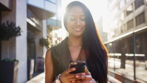 Attractive young woman smiling as she uses her phone walking down the street in  Stock Photos