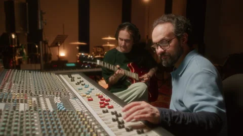 Audio Engineer Adjusting Mixing Console while Musician Playing Guitar in Studio Stock Footage 334369974