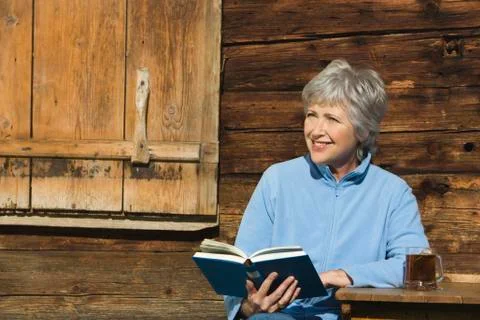 Austria, Senior woman reading book by log cabin, portrait Stock Photos