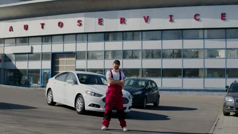 Auto engineer in uniform stands by car looks into camera crosses arms at auto Stock Footage 276092064