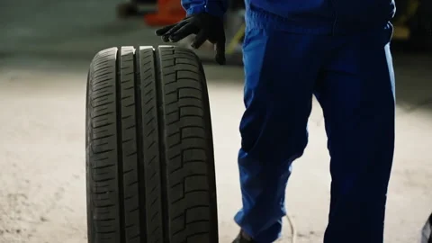 Auto mechanic in blue uniform and protective gloves rolling a car tire across a Stock Footage 333839308