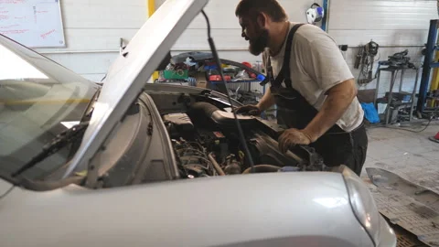 Auto mechanic in uniform looking under hood of vehicle at garage Stock Footage 328641086