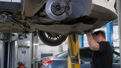 An auto mechanic working under a car on a car lift in a garage. Auto repair shop Stock Footage 337221067