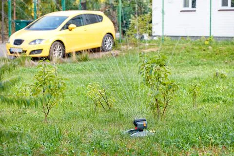 An automatic sprayer waters a young garden and lawn in the backyard of a resi Stock Photos